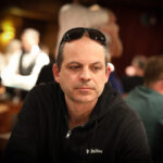 Ein Mann in einem dunklen Anzug und einer roten Krawatte spielt entschlossen Poker mit ernstem Gesichtsausdruck an einem eleganten Tisch mit grünem Filz und Chips.