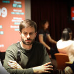 Ein Mann sitzt an einem Pokertisch und hält eine Hand mit Spielkarten. Er hat einen konzentrierten Gesichtsausdruck und trägt eine dunkle Brille. Im Hintergrund sind weitere Personen und Chips