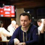 Ein Mann in einem schwarzen Anzug und einer schwarzen Sonnenbrille sitzt an einem Pokertisch und hält zwei Spielkarten in der Hand. Er wirkt konzentriert und ernsthaft, während er auf seine