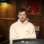 Ein Mann in einem schwarzen Anzug und einer Sonnenbrille sitzt an einem Pokertisch und hält zwei Karten in der Hand. Er schaut konzentriert auf seine Karten, während im Hintergrund andere Spieler
