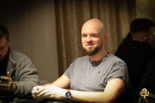Ein Mann in einem schwarzen Anzug sitzt an einem Pokertisch und betrachtet konzentriert seine Spielkarten. Um ihn herum sind weitere Spieler und Pokerchips zu sehen.