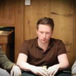 Ein Mann in einem schwarzen Anzug sitzt an einem Pokertisch und hält zwei Spielkarten in der Hand. Er schaut konzentriert auf die Karten. Im Hintergrund sind Pokerchips und andere Spieler zu