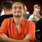 Ein Mann mit schwarzer Sonnenbrille sitzt an einem Pokertisch und hält seine Karten in der Hand, während er konzentriert auf seine Gegner schaut.