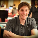 Ein Mann in einem Anzug sitzt an einem Pokertisch und zeigt eine Siegerhand mit einem breiten Lächeln. Andere Spieler sitzen um den Tisch herum und schauen gespannt zu.