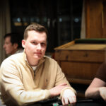 Ein Mann und eine Frau sitzen an einem Pokertisch, beide konzentriert auf ihre Karten und Chips. Der Mann trägt einen Anzug und die Frau ein elegantes Kleid. Im Hintergrund sind weitere Pokertische