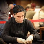 Ein professioneller Pokerspieler mit Sonnenbrille und Kopfhörern sitzt an einem Pokertisch mit Chips und Karten.