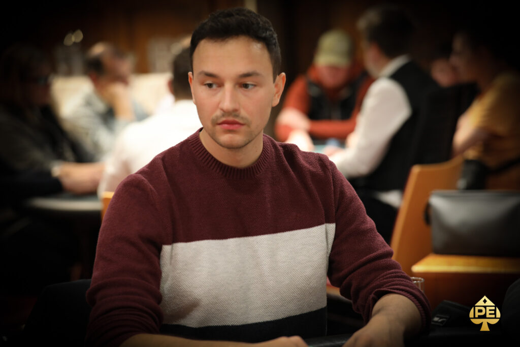 Ein Mann in einem dunkelblauen Anzug und einer Fliege steht lächelnd vor einem Pokertisch mit Spielkarten und Pokerchips. Im Hintergrund sind weitere Personen und eine Casino-Atmosphäre zu