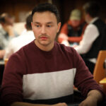 Ein Mann in einem dunkelblauen Anzug und einer Fliege steht lächelnd vor einem Pokertisch mit Spielkarten und Pokerchips. Im Hintergrund sind weitere Personen und eine Casino-Atmosphäre zu