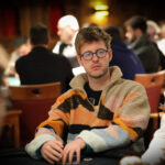 Ein Mann sitzt an einem Pokertisch mit Chips und Karten vor sich. Er hat eine konzentrierte Miene und trägt eine schwarze Kappe.