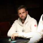 Ein Mann in einem schwarzen Anzug sitzt an einem Pokertisch mit grünem Filz und hält zwei Spielkarten in der Hand. In der Mitte des Tisches liegen weitere Karten und Pokerchips. Im