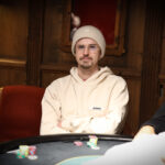 Ein Mann in einem Anzug und mit einer Sonnenbrille sitzt an einem Pokertisch und hält Pokerchips in der Hand. Andere Spieler und Karten sind auf dem Tisch zu sehen.