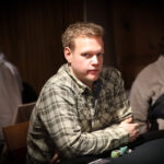 Ein Mann und eine Frau sitzen an einem Pokertisch und spielen eine Partie Poker. Der Mann hat eine Hand mit Karten in der Hand, während die Frau konzentriert auf ihre eigenen Karten schaut. Im Hintergrund
