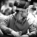 Ein Mann sitzt an einem Pokertisch und hält zwei Karten in der Hand, während er konzentriert auf die Gemeinschaftskarten in der Mitte des Tisches schaut. Andere Spieler und Dealer sind im H