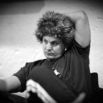 Ein Mann und eine Frau sitzen an einem Pokertisch und betrachten konzentriert ihre Karten. In der Mitte des Tisches liegen Pokerchips und Spielkarten. Im Hintergrund sind weitere Spieler und eine bele