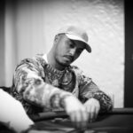 Ein eleganter Mann in einem schwarzen Anzug und einer dunkelblauen Krawatte sitzt an einem Pokertisch und hält seine Spielkarten in der Hand. Im Hintergrund sind unscharfe Spieler und Pokerchips