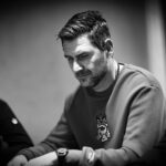 Ein Mann in einem dunkelblauen Anzug und einer blauen Krawatte steht vor einem Pokertisch mit Chips und Karten. Er hält zwei Spielkarten in der Hand und schaut konzentriert auf den Tisch