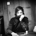 Ein Mann in einem schwarzen Anzug und einer schwarzen Sonnenbrille sitzt an einem Pokertisch und hält seine Karten. Er wirkt konzentriert und selbstbewusst. Im Hintergrund sind andere Pok