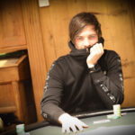Ein Mann in einem schwarzen Anzug und einer roten Krawatte sitzt an einem Pokertisch und hält seine Spielkarten in der Hand. Im Hintergrund sind Casino-Chips und andere Spieler verschwommen zu sehen.