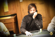 Ein Mann in einem schwarzen Anzug und einer roten Krawatte sitzt an einem Pokertisch und hält seine Spielkarten in der Hand. Im Hintergrund sind Casino-Chips und andere Spieler verschwommen zu sehen.