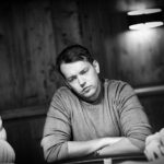 Ein professioneller Pokerspieler in einem schwarzen Anzug und mit Sonnenbrille, der konzentriert Karten auf einem Pokertisch betrachtet. Im Hintergrund sind andere Spieler und grüne Pokertisch