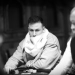 Ein Mann in einem dunkelblauen Anzug und einer hellblauen Krawatte sitzt an einem Pokertisch mit grünem Filz. Er hält Karten in der Hand und schaut konzentriert auf den T