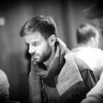 Ein Mann sitzt an einem Pokertisch und betrachtet konzentriert seine Karten. Um ihn herum sind weitere Spieler zu sehen, die ebenfalls ihre Karten prüfen. Im Hintergrund sind Pokerchips und Get