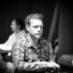 Ein Mann sitzt an einem Pokertisch und hält zwei Spielkarten in der Hand. Er hat eine konzentrierte Miene und schaut auf seine Karten. Im Hintergrund sind Chips und Spielkarten auf dem T