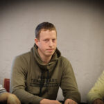 Ein Mann in einem schwarzen Anzug sitzt an einem Pokertisch und hält zwei Spielkarten in der Hand. Er scheint konzentriert und selbstbewusst zu sein, während er an einem Pokerspiel teilnim