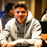 Ein Mann in einem dunkelblauen Anzug sitzt an einem Pokertisch und hält eine Hand mit Spielkarten. Er wirkt konzentriert und fokussiert. Die anderen Spieler am Tisch sind unscharf
