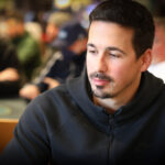 Ein Mann sitzt an einem Pokertisch und hält Karten in der Hand, während er konzentriert auf seine Gegner schaut. Im Hintergrund sind weitere Spieler und Chips zu sehen.