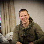 Ein Mann in einem schwarzen Anzug sitzt an einem Pokertisch und hält seine Karten hoch, während er konzentriert auf seine Gegner schaut. Die Chips und Karten auf dem Tisch deuten auf ein intens