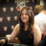 Ein professioneller Pokerspieler mit Sonnenbrille und Kappe sitzt an einem Pokertisch und betrachtet konzentriert seine Karten. Im Hintergrund sind Chips und Spielkarten zu sehen.