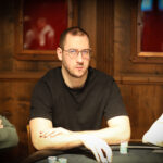 Ein Mann in einem Anzug und mit einer Sonnenbrille spielt Poker an einem Tisch mit Karten und Chips. Er hat eine ernste Miene und betrachtet konzentriert seine Karten. Andere Spieler sind im