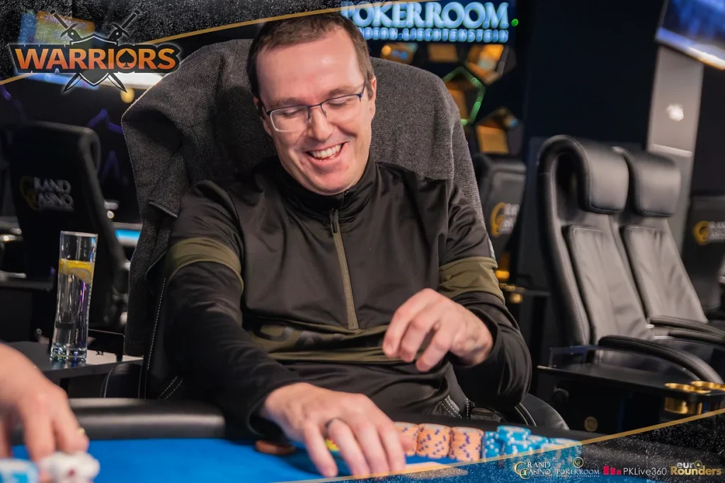 Ein Mann in einem schwarzen Anzug und einer Sonnenbrille hält zwei Spielkarten in seiner Hand und schaut ernst in die Kamera, während er in einem luxuriösen Pokerraum sitzt.