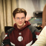 Ein Mann mit dunklen Haaren und einem dunklen Anzug sitzt lächelnd an einem Pokertisch und hält Karten in der Hand.