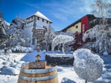Ein Mann in winterlicher Kleidung und mit einer Mütze steht neben einem Schneemann in einem verschneiten Park. Im Hintergrund sind Bäume mit Schnee bedeckt.