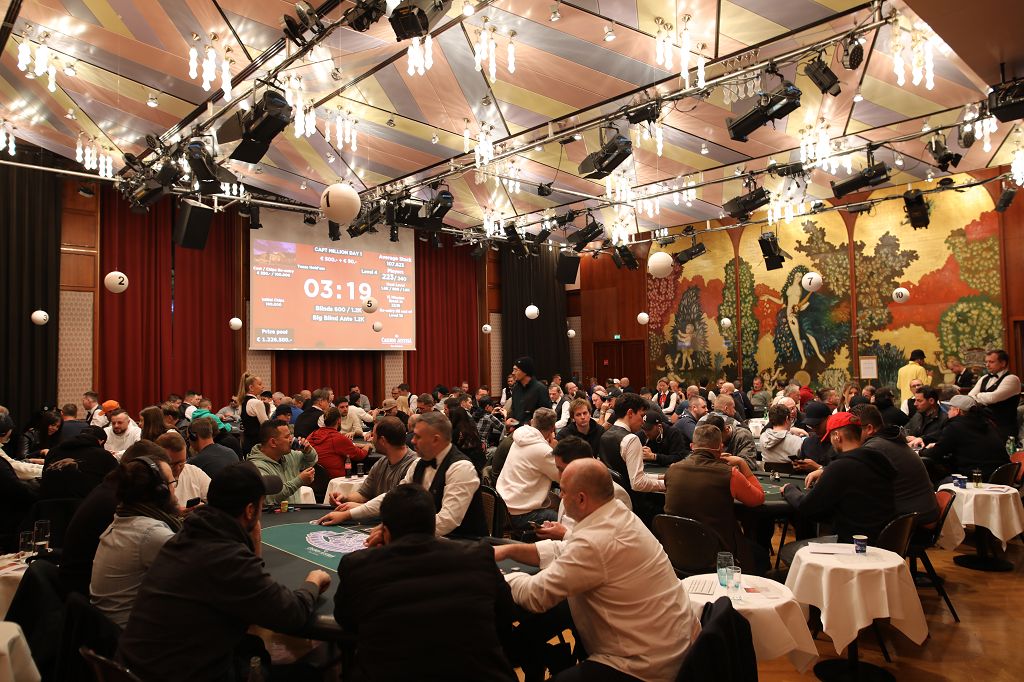 Ein Gruppenfoto von Pokerspielern, die an einem Turnier teilnehmen. Sie sitzen an einem Tisch mit Pokerchips und Karten vor sich. Die Spieler sind konzentriert und in Aktion.