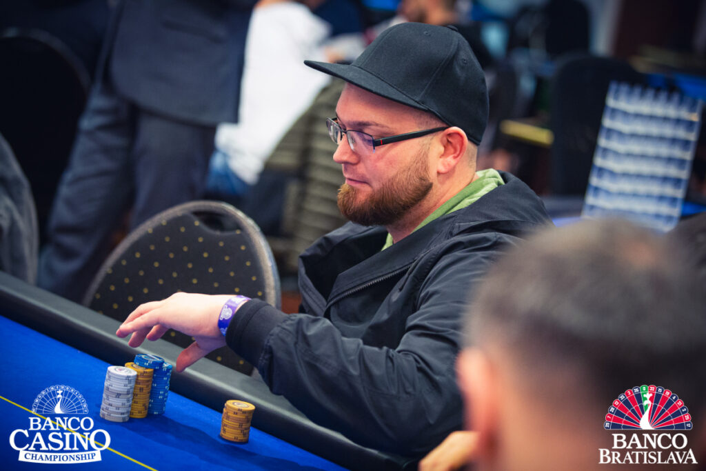 Ein Pokertisch mit Stapeln von Pokerchips in verschiedenen Farben, wobei ein Spieler mit einem großen Stapel von blauen Chips der Chipleader ist. Andere Spieler und Karten sind im Hintergrund zu sehen.