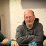 Ein Mann mittleren Alters mit braunen Haaren und einem dunkelblauen Hemd sitzt entspannt an einem Pokertisch und betrachtet konzentriert seine Karten.