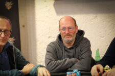 Ein Mann mittleren Alters mit braunen Haaren und einem dunkelblauen Hemd sitzt entspannt an einem Pokertisch und betrachtet konzentriert seine Karten.