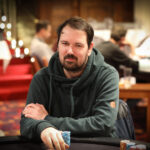 Ein Mann mit dunklen Haaren und einem Bart, der ein blaues Hemd trägt und vor einem Pokertisch sitzt. Er hält Karten in der Hand und schaut konzentriert auf seinen Gegner.