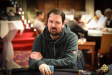 Ein Mann mit dunklen Haaren und einem Bart, der ein blaues Hemd trägt und vor einem Pokertisch sitzt. Er hält Karten in der Hand und schaut konzentriert auf seinen Gegner.