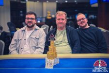 Ein Mann sitzt an einem Pokertisch mit Chips und Karten vor sich. Er hat eine ernste Miene und trägt eine dunkle Brille. Im Hintergrund sind weitere Spieler und eine elegante Raumausstattung zu sehen