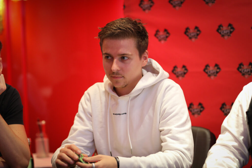 Ein junger Mann in einem dunklen Anzug und einer weißen Hemdweste sitzt an einem Pokertisch und betrachtet konzentriert seine Karten. Er hat dunkles Haar und trägt eine Brille.
