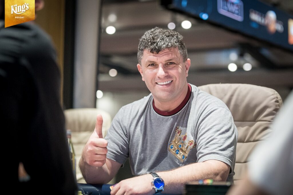Ein Mann in einem dunklen Anzug sitzt an einem Pokertisch und hält einige Spielkarten in der Hand. Im Hintergrund sind andere Spieler und Chips auf dem Tisch zu sehen.
