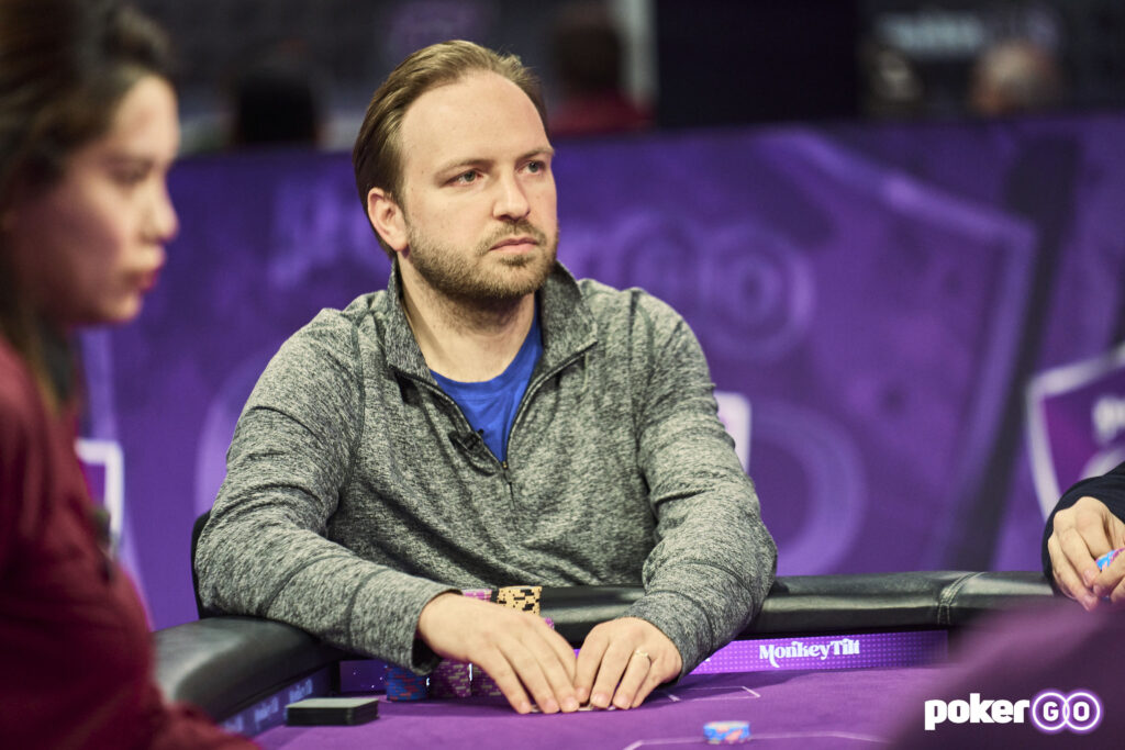 Ein Mann sitzt an einem Pokertisch während eines Turniers. Er trägt eine Brille, ein schwarzes Hemd und hat einen konzentrierten Blick auf seine Karten. Im Hintergrund sind weitere Spieler und eine Dealerin
