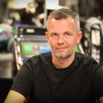Ein Mann sitzt entspannt an einem Pokertisch und betrachtet seine Karten. Er trägt eine dunkle Brille und einen schwarzen Anzug. Im Hintergrund sind Pokerchips und ein Getränk zu