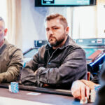 Ein Mann sitzt an einem Pokertisch und schaut konzentriert auf seine Karten. Er hat dunkles Haar, trägt eine Brille und ein dunkles Hemd. Im Hintergrund sind Chips und Spielkarten