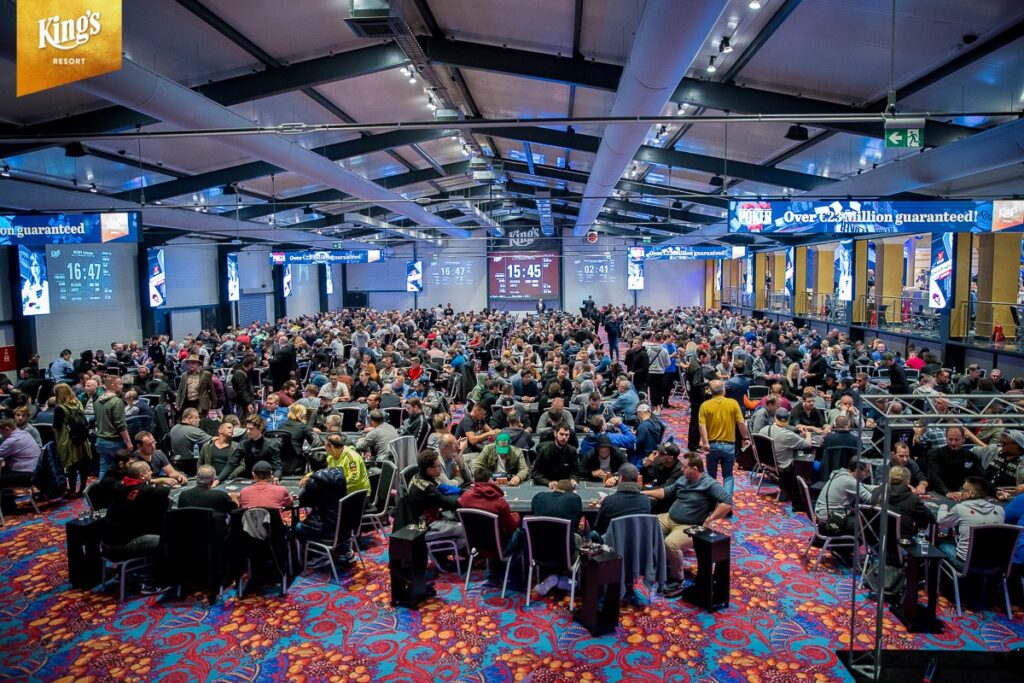 Ein vollbesetzter Pokertisch in einer eleganten Pokerrunde, bei der Spieler mit Pokerchips und Karten spielen. Die Spieler sind konzentriert und fokussiert auf ihre Hände. Helle Beleucht