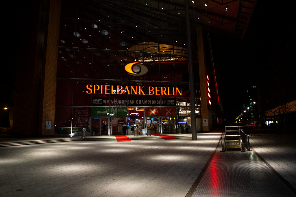 Ein eleganter Eingangsbereich einer Spielbank in Berlin mit einer großen, gläsernen Eingangstür, rotem Teppich, Kronleuchter und stilvoller Einrichtung.